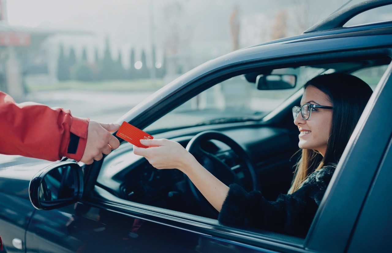 Kobieta kierująca samochodem przekazuje kartę lojalnościową ORLEN w Portfelu pracownikowi stacji.
