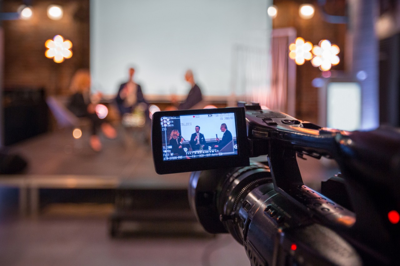 Businesswoman and businessmen discussing during online seminar, sitting on armchairs on the stage. Focus on video camera.