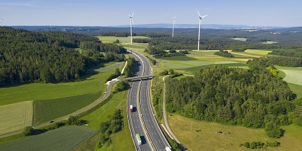 Stacje paliw ORLEN. Autostrada z poruszającymi się ciężarówkami, a w tle rozmieszczone turbiny wiatrowe, generujące czystą energię.