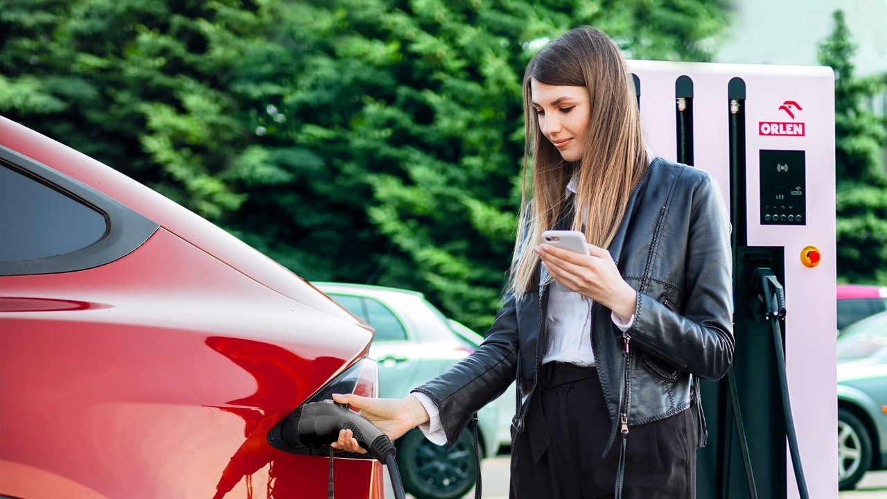  ORLEN Charge aplikacja. Kobieta trzyma w dłoniach telefon komórkowy oraz ładuje samochód elektryczny na stacji paliw. 
