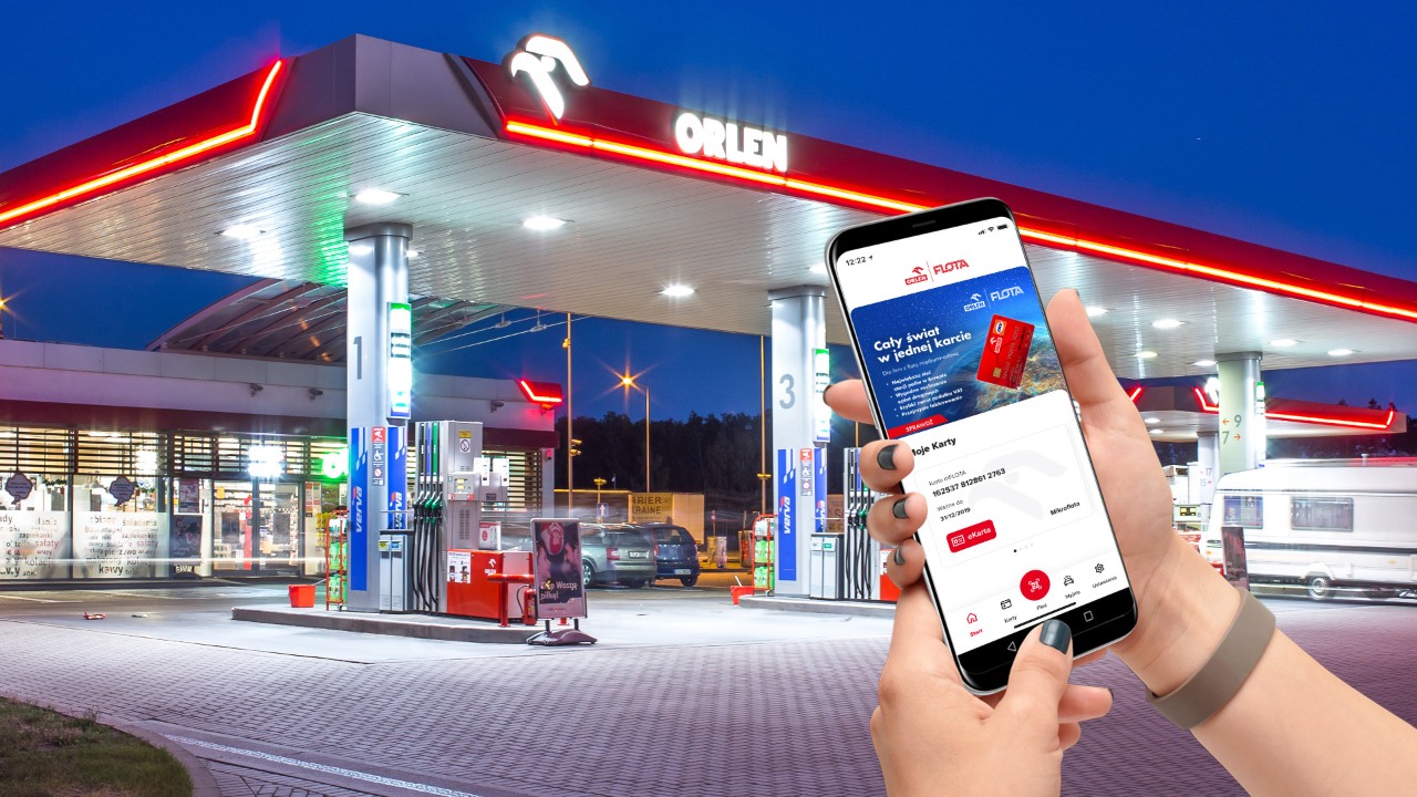  mFLOTA ORLEN aplikacja. Kobieta trzyma w dłoniach telefon komórkowy, na którym wyświetla się aplikacja mFlota ORLEN. W tle stacja paliw ORLEN. 