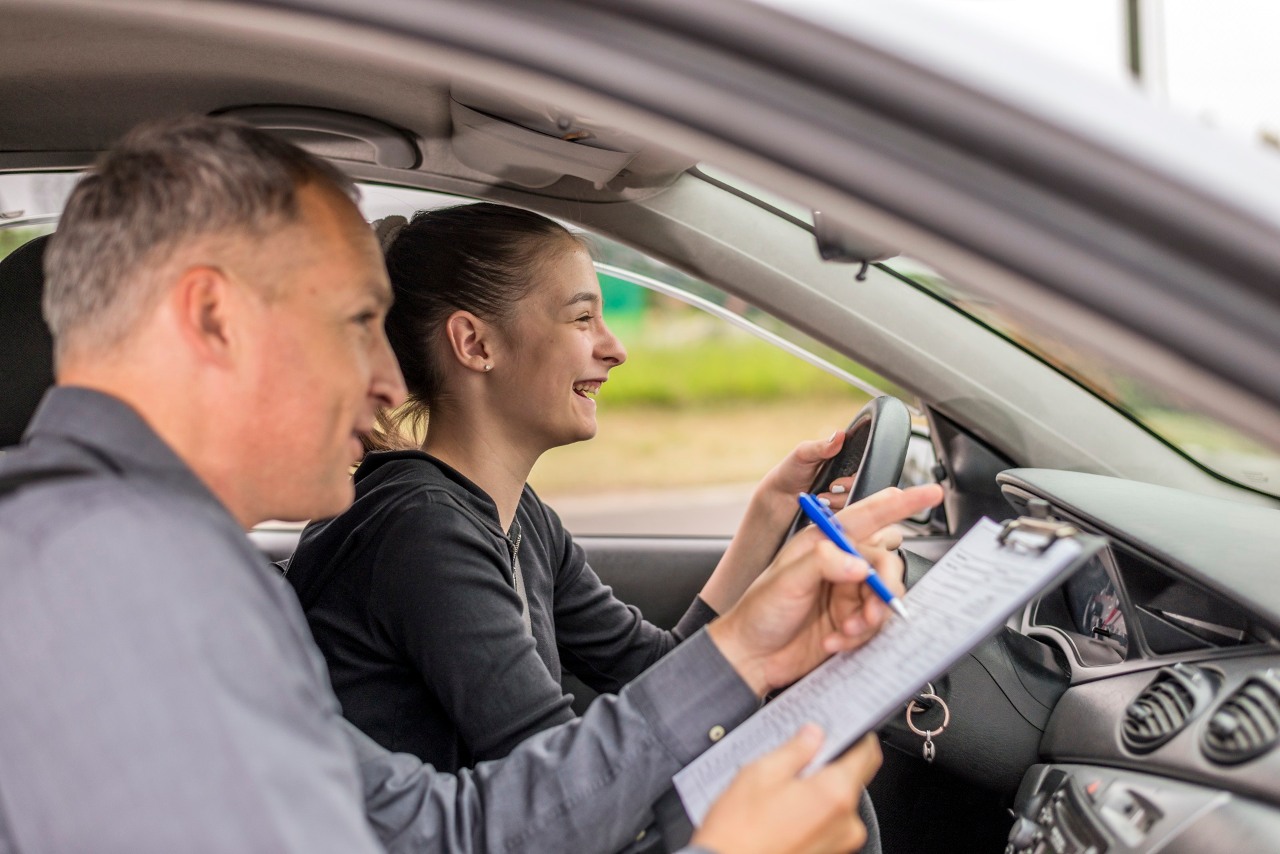 Driving male instructor  taking notes while holding clipboard while sitting next to driving female teen student learning to drive during bright suuny day.