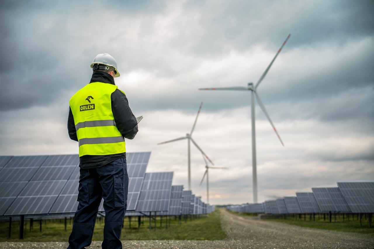 Energia odnawialna, energia wiatrowa i energia słoneczna – Grupa ORLEN. Pracownik firmy ORLEN podczas prac na terenie farmy fotowoltaicznej. W tle turbiny wiatrowe.  